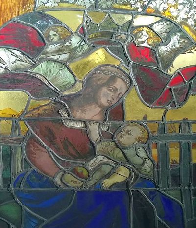 Ein in einen Holzrahmen gefasstes Glasfenster zeigt Maria mit dem Jesuskind. Über ihr schweben zwei Engel, die eine Krone über Marien Kopf halten.