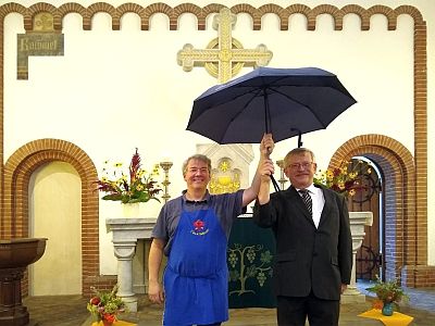 Zwei Männer stehen vor dem Altar einer Kirche und halten einen Regenschirm über sich. Sie lachen. Hinter ihnen überragt sie das Altarkreuz.