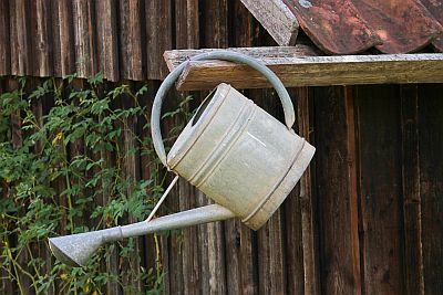 Eine Gießkanne aus Zinkblech hängt an der hölzernen Dachrinne eines Gartenhauses.