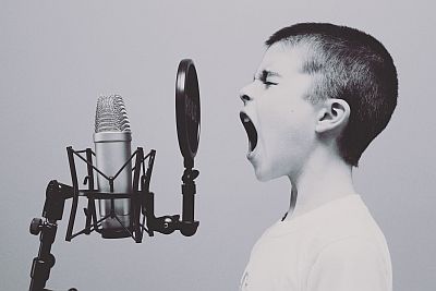 Ein Junge steht mit offenem Mund, angestrengtem Gesichtsausdruck und geschlossenen Augen vor einem Studiomikrofon mit Poppschutz. Es sieht nicht nach Gesang aus, eher nach einem Schrei, aber man kann nicht ganz sicher sein.
