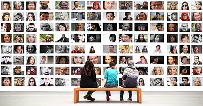 An einer Wand hängen über 100 Fotos mit Gesichtern von Menschen aus allen Teilen der Welt. Auf einer Bank davor sitzen drei auch sehr verschiedene Menschen und tauschen sich über die Bilder aus. Einige Menschen sind mehrmals abgebildet. Soll uns das etwas sagen? Wie auch immer: es handelt sich um Kunst.