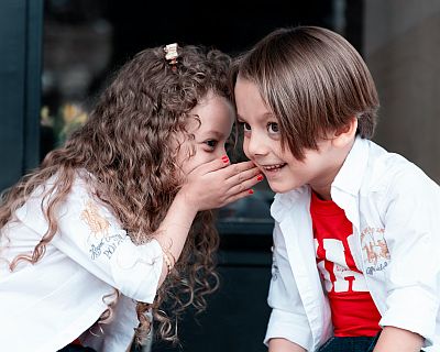 Zwei Kinder tuscheln miteinander. Hinter vorgehaltener Hand sagt ein Mädchen einem Jungen etwas. Er hört aufmerksam hin, seine Augen leuchten, sein Mund ist geöffnet.