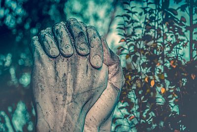 Wir sehen als Teil einer Statue zwei im Gebet gefaltete, ringende Hände. Im Hintergrund sind Pflanzen eines Gartens oder Friedhofs erkennbar.