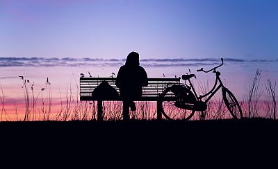 Eine Frau sitzt auf einer Bank, den Rucksack neben sich, das Fahrrad steht dabei, ein Damenrad, eine herrliche 'Holländermühle'. Wir sehen Frau, Bank und Rad nur von hinten, ihre Konturen erscheinen vor dem Himmel über der untergegangenen Sonne.