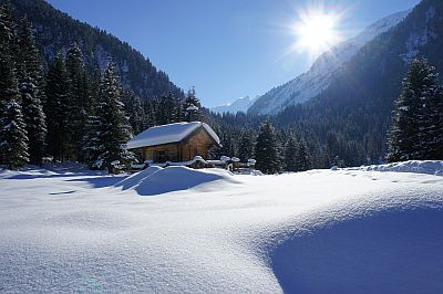 Wir sehen eine Winterlandschaft im gebirge. Die tiefstehende Sonne leuchtet auf den weißen und tiefen Schnee. In der Mitte im Tal steht eine Kapelle.