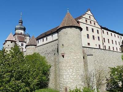 Wir schauen von unten auf eine Festungsanlage. Vor uns erhebt sich an der Ecke der Festungsmauer ein mächtiger Wehrturm. Im Inneren der Festung sind ein Schloss und ein Kirchturm erkennbar.
