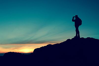 Ein Wanderer steht auf dem Gipfel eines Berges und Blickt der Sonne entgegen, die gerade in weiter Ferne über dem Horizont aufgeht. Es ist leicht bewölkt, der Himmel ist noch frisch blau und es ist kühl.