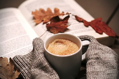 Es wird gemütlich. Neben einer Tasse mit frischem Kaffee liegen ein Buch und gestrickte Pulswärmer. Auf dem Buch liegen eine bunte Eichenblätter, es ist Herbst.