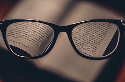 Eine Lesebrille wird bei gedämpftem Licht vor ein Buch gehalten. Hinter ihren Gläsern ist die kyrillische Schrift des Textes klar zu erkennen. Um die Brille herum ist alles verschwommen.