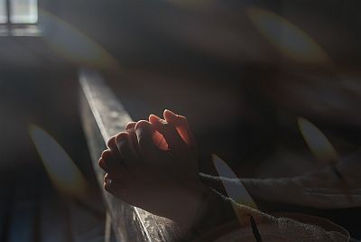 Aus einem Fenster im Hintergrund fällt Licht auf die Hände einer jungen Frau, die sie zum Gebet gefaltet auf die Kirchenbank gelegt hat.