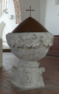 Foto: ein zirka 1 Meter hoher kelchförmiger Taufstein aus Stein. Auf seiner runden Öffnung sitzt eine kegelförmige bronzene Haube mit einem schlichten Kreuz.