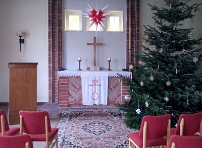 Abbildung: Der Blick fällt auf einen Altar, über dem ein Herrnhuter Stern hängt. Links steht ein Lektorenpult, rechts ein Weihnachtsbaum. Im Vordergrund sind leere Stühle zum Altar ausgerichtet.