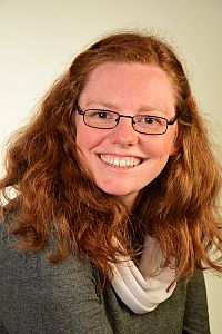 Abbildung: eine junge Frau mit langen roten Haaren und einer Brille lächelt uns an.  Es ist unsere Pfarrerin Elisabeth Koppehl.
